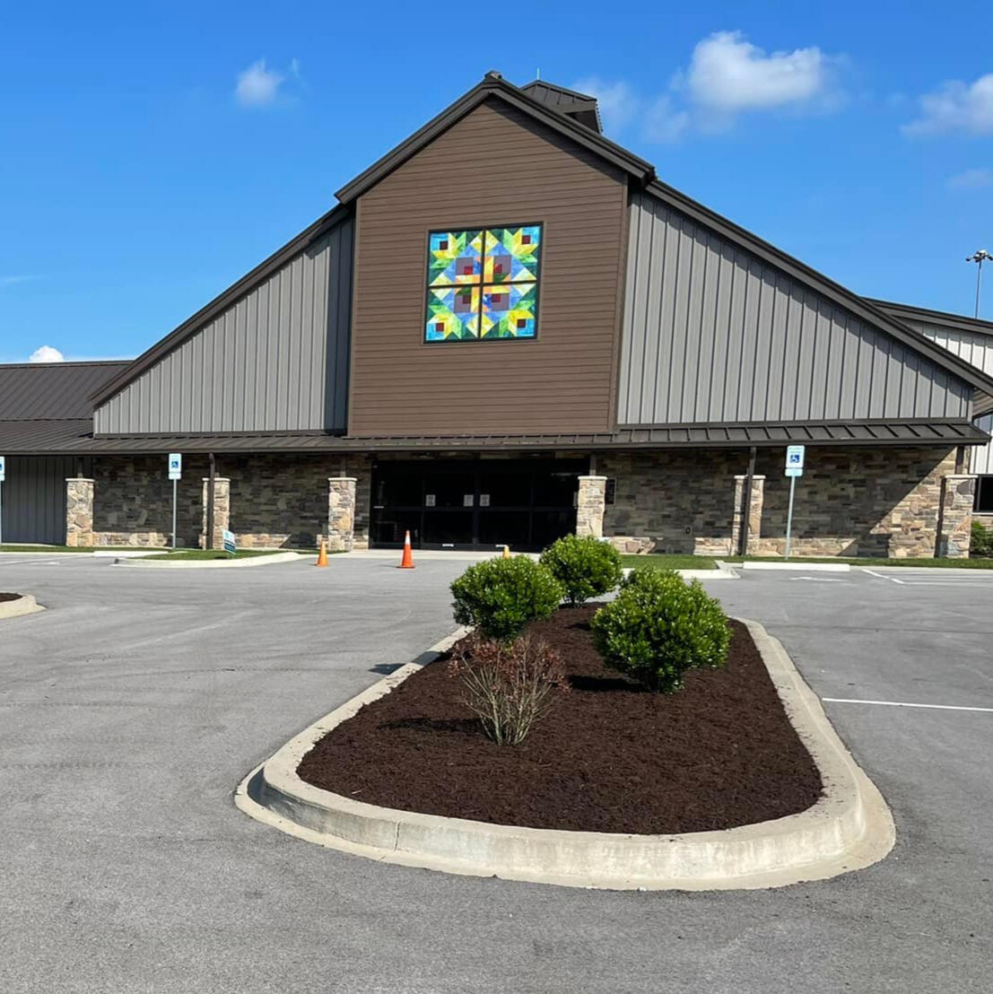 Local Church A median of fresh, dark brown mulch with 3 freshly-trimmed shrubs in front of a large, brown building with pointed roof. Large, stained glass window immediately below the apex of the roof.