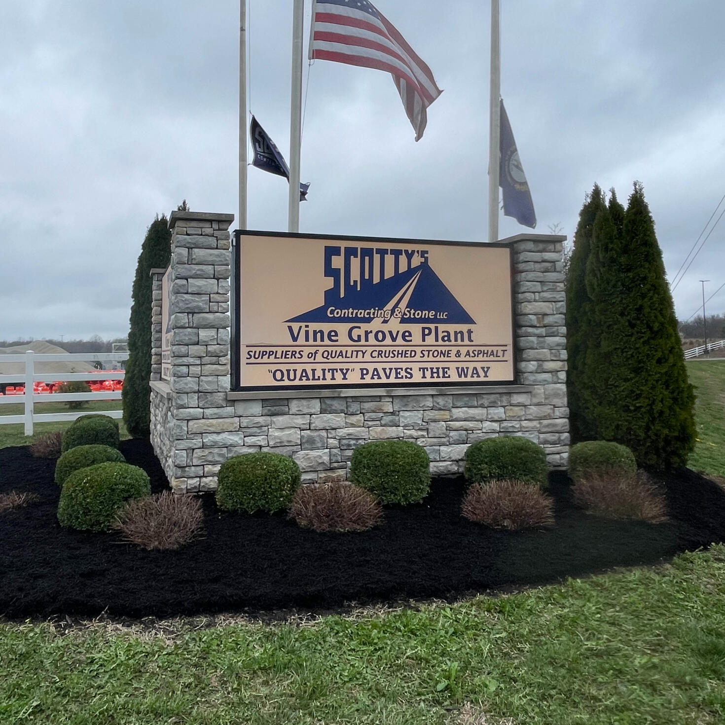 Scotty's Vine Grove Plant Fresh, black mulch and freshly-trimmed shrubs and pointed pine trees surrounding a brick sign that reads 'Scott's Vine Grove Plant'. Three visible flags including the American flag and Kentucky state flag.
