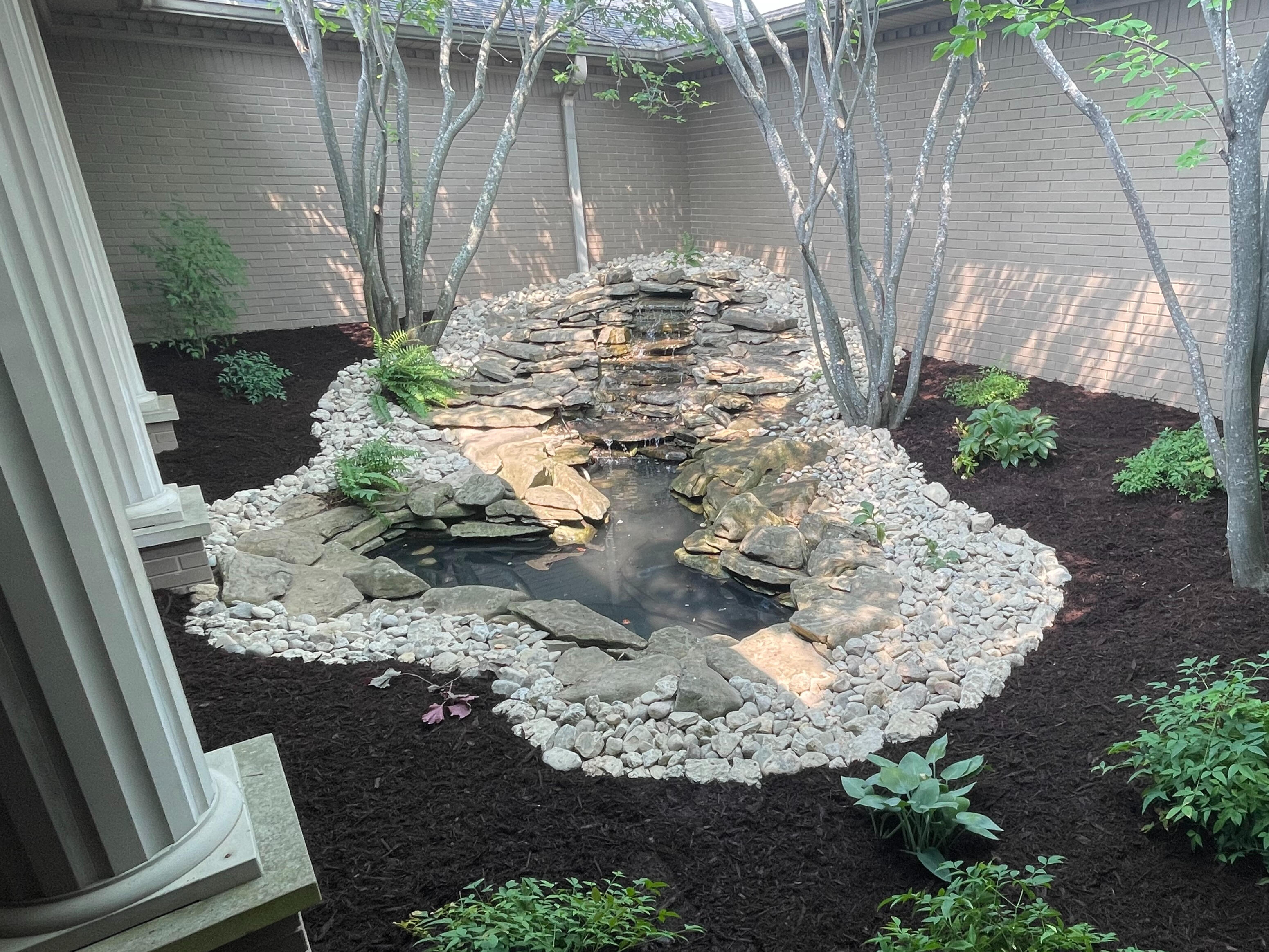 Rock Fountain A medium-sized rock and stone with water trickling downward. A few small plants and dark mulch surround the fountain.