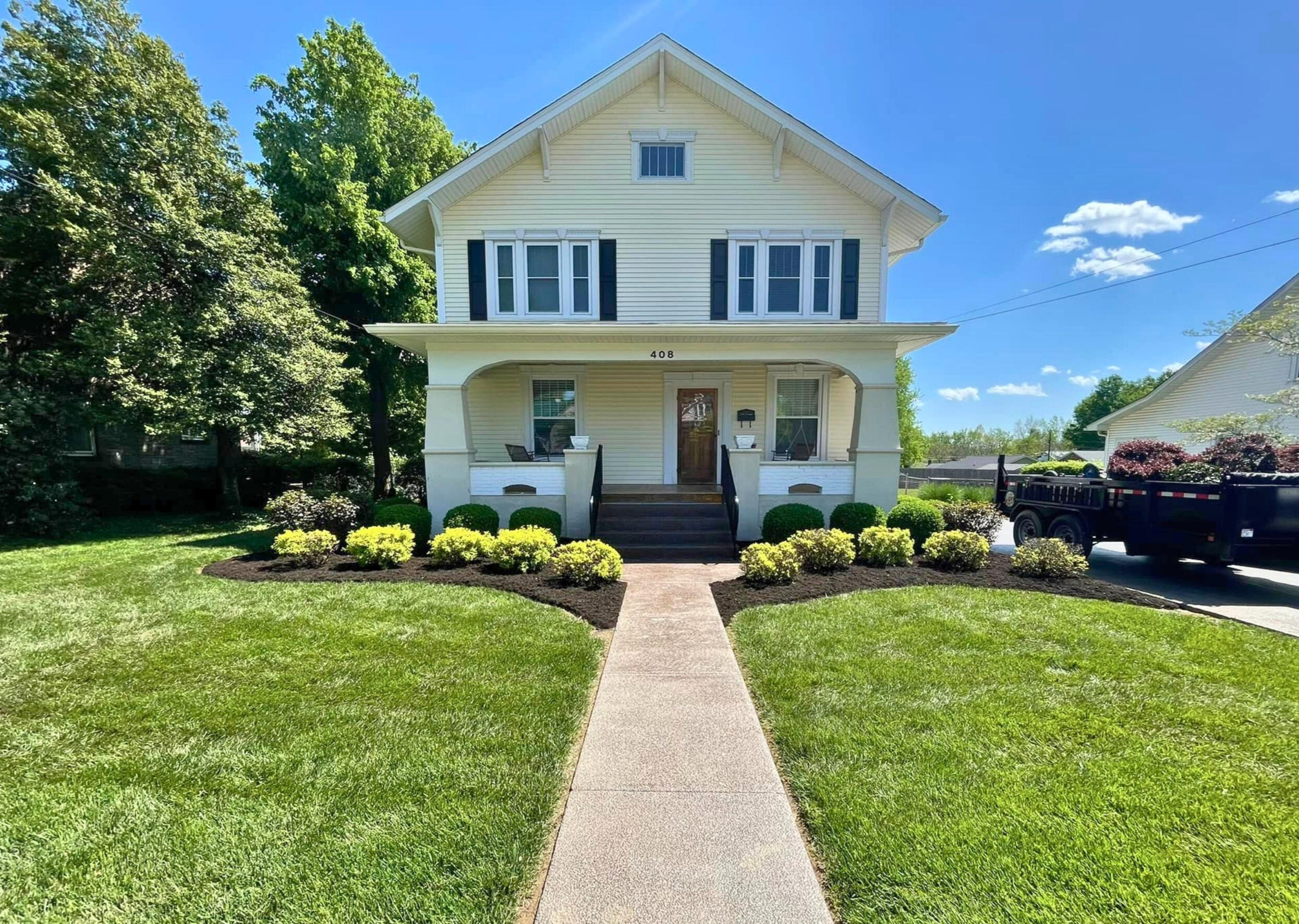 Residential Landscaping A freshly moved lawn with fresh, dark brown mulch and small shrubs in front of an older, two-story home with light yellow siding and brown wood door on a partly cloudy day.